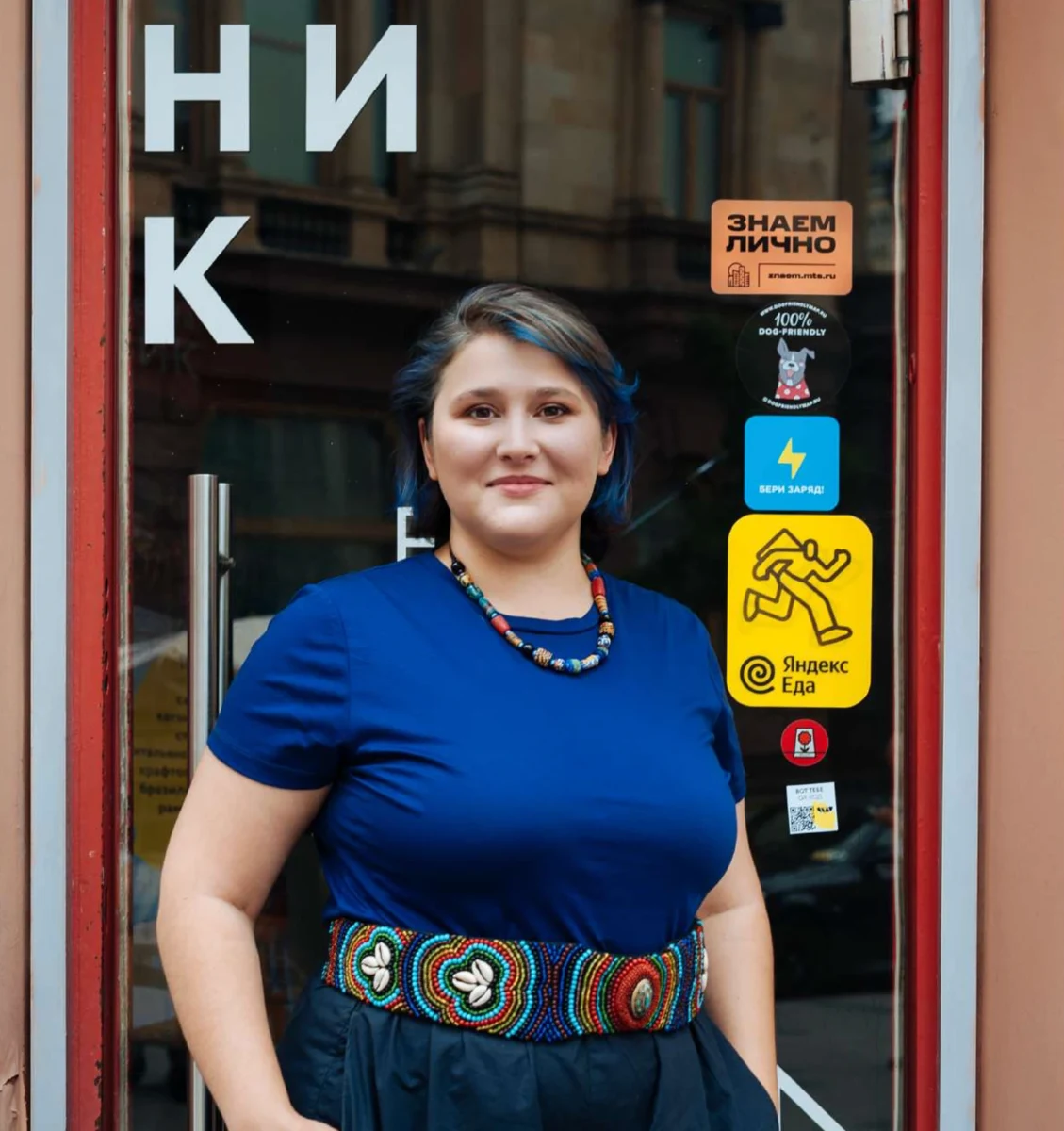 A business owner at the door of her shop, with a Knowing in Person sticker on the glass